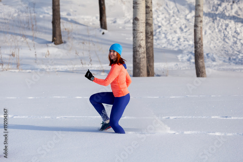 Young woman in a bright blue hat, orange sweater runnnig on a snow on a bright winter day in forrest. Jogging in winter forest
