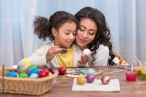 African American mother hugging daughter while she paints easter eggs