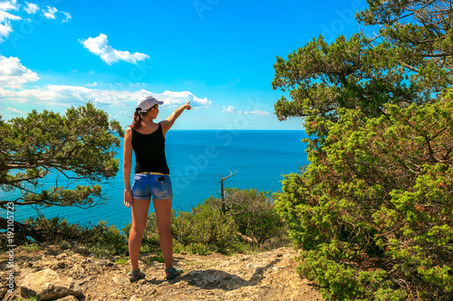 A young tourist girl stands on the edge of a mountain and looks away at the beautiful summer seascape at a resort in Crimea