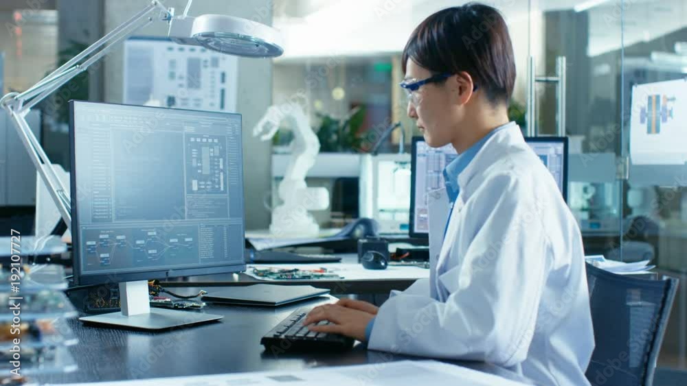 Asian Scientist Sitting at His Desk Doing Sophisticated Coding and Programming on His Desktop Computer. In the Background Computer Science Research Laboratory with Robotic Arm Model.