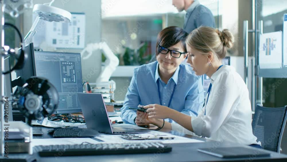 Asian Scientist Sitting at His Desk Consults Senior Engineer They Connect Printed Circuit Board to a Laptop and Start Coding. In the Background Computer Science Research Laboratory with Robotic Arm.
