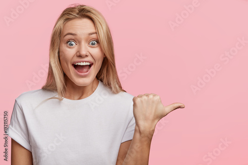 Attractive overjoyed female dressed casually, expresses surprisment and excitedness, indicates at blank copy space for your advertising content, isolated over pink background. Startled woman photo
