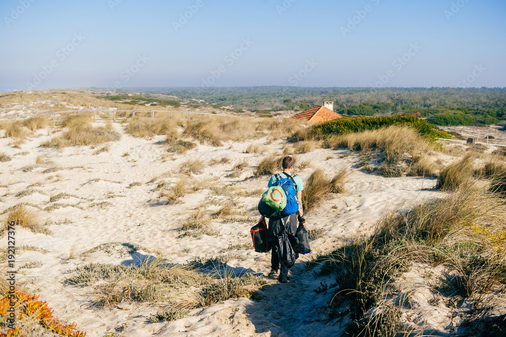 Fototapeta premium Homeless hermit carrying many bags with belongings in sandy dunes beyond sea in sunny day. Man from behind travelling alone. Unknown male with backpacks and heavy buggage in hands hiking outdoor.