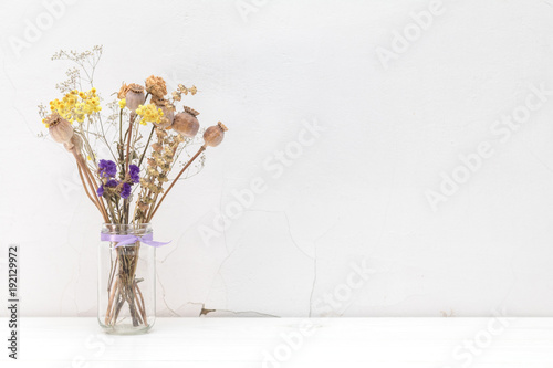 Dried flowers and poppy heads in a glass jar on white cracked wall background.