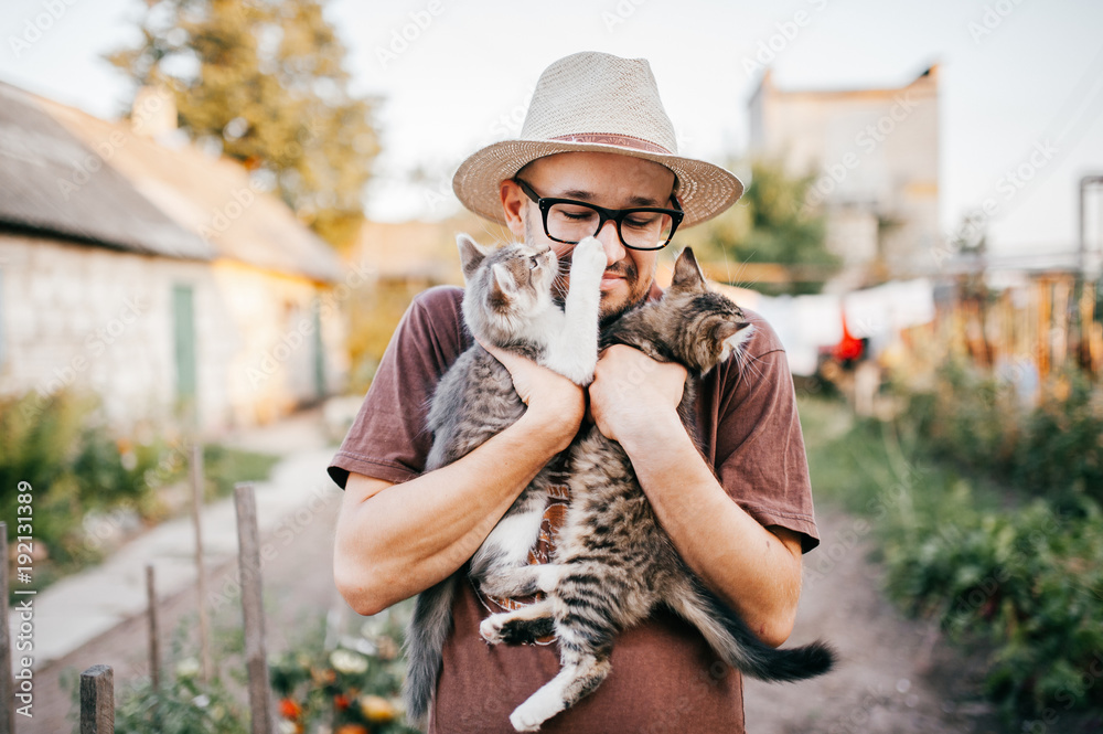 Φωτογραφία Happpy young bearded farmer holding two little kitten in hands outdoor in village with abstract background