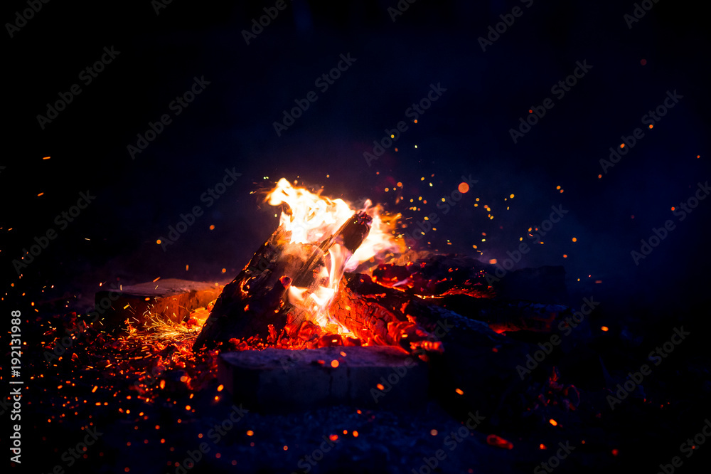 Burning wood at night. Campfire at touristic camp at nature in mountains. Flame amd fire sparks on dark abstract background. Cooking barbecue outdoor. Hellish fire element. Fuel, power and energy.