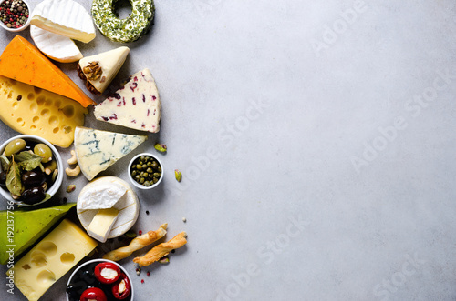 Assortment of hard, semi-soft and soft cheeses with olives, grissini bread sticks, capers, grape, on grey concrete backgound. Top view, copy space, flat lay. Cheese selection appetizer plate.