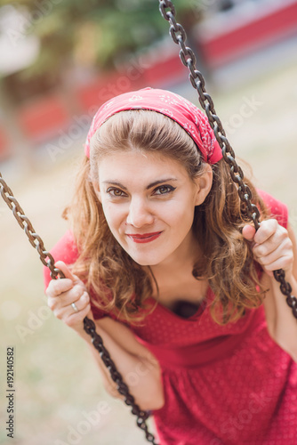 Happy young woman in red retro dress swinging on a swing in a park on a sunny day