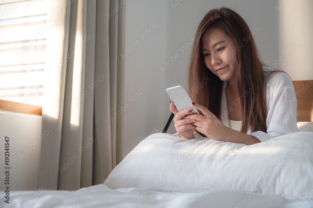 Portrait image of a beautiful Asian woman sitting on the bed , using and looking at mobile phone after woke up with feeling happy Portrait image of a beautiful Asian woman sitting on the bed , using and looking at mobile phone after woke up with feeling happy