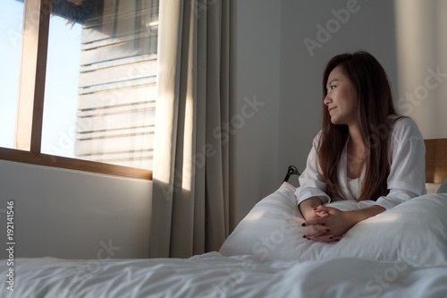 Portrait image of a beautiful Asian woman sitting on the bed and looking outside the window with feeling happy and relaxed