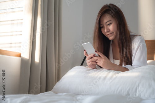 Portrait image of a beautiful Asian woman sitting on the bed , using and looking at mobile phone after woke up with feeling happy