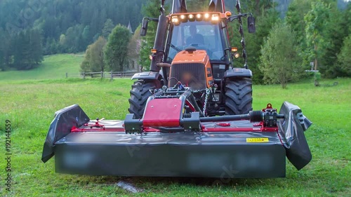 A tractor is standing still in the middle of the grass field and the farmer presses a button on the vehicle and the grass cutting machinery starts lowering down.