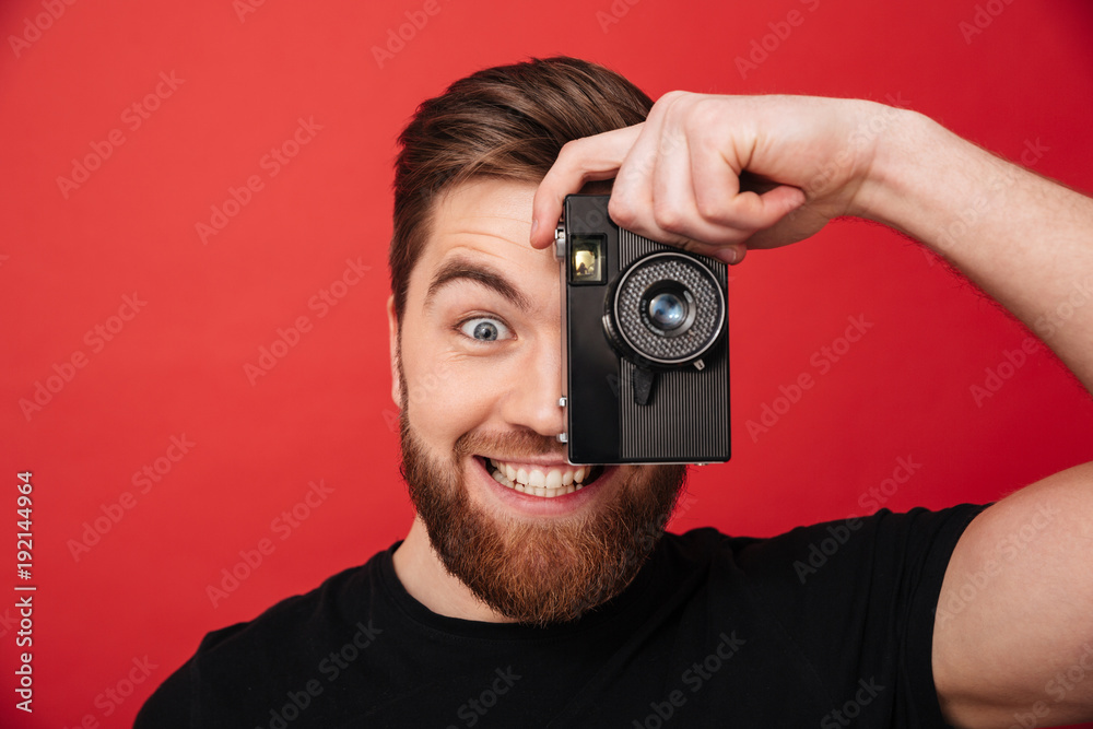 Portrait of smiling amateur male photographer making pictures using retro camera, isolated over red background - obrazy, fototapety, plakaty 