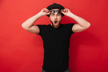 Portrait of surprised stylish man 30s in hat posing on camera and taking off black sunglasses, isolated over red background
