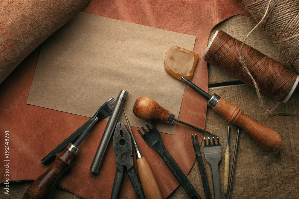Naklejka premium Set of leather craft tools on wooden background. Workplace for shoemaker. Piece of hide and working handmade tools on a work table.