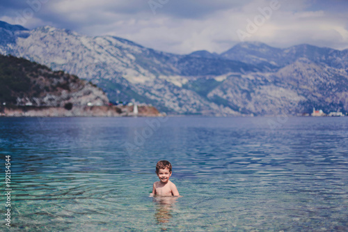 Little smiling boy in the sea with blue and turquoise water. In the background blue mountains.