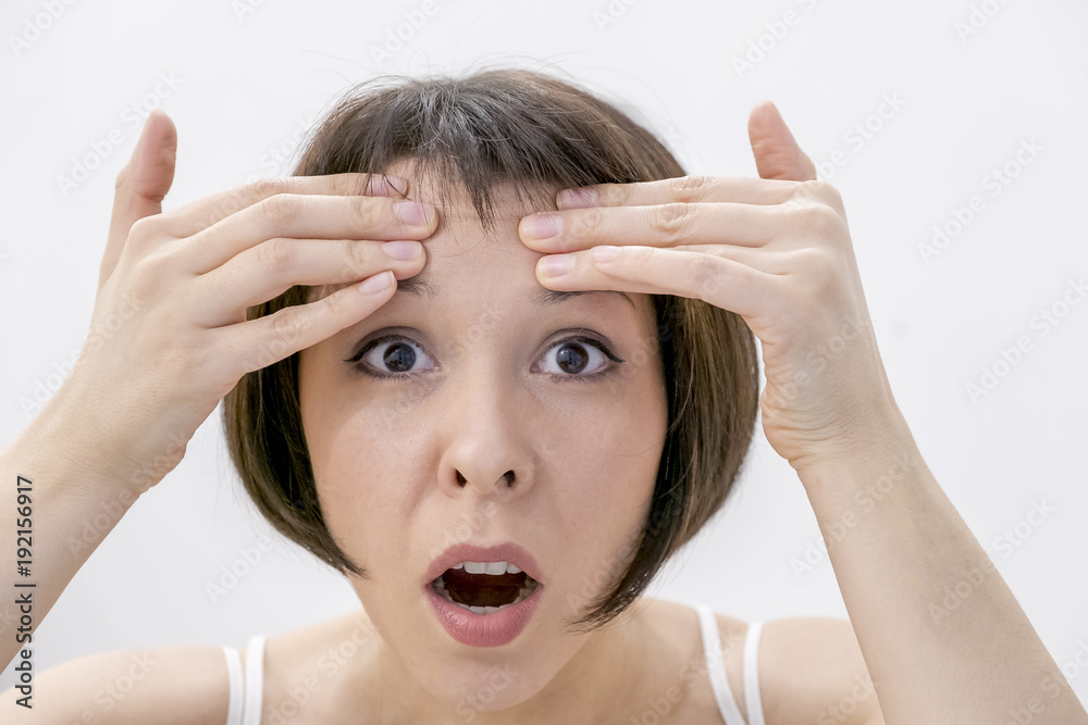 A young woman checking wrinkles on her forehead. Horrified young woman looking in the bathroom mirror staring open mouthed a first sign of ageing A young woman checking wrinkles on her forehead. Horrified young woman looking in the bathroom mirror staring open mouthed a first sign of ageing