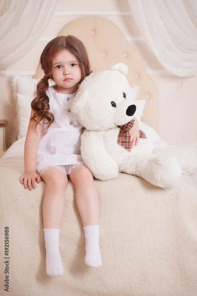 A little girl in her room sits on a bed and holds a bear. A child in a white dress with a toy. Beige interior. A little girl in her room sits on a bed and holds a bear. A child in a white dress with a toy. Beige interior.