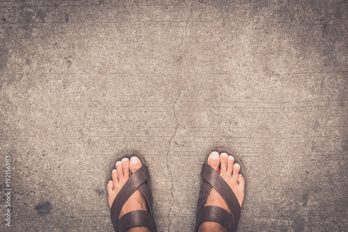 man feet wearing some brown flip flops standing on the asphalt concrete floor