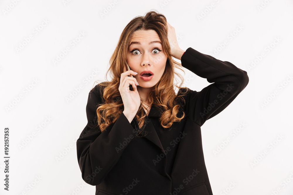 Picture of attractive woman with long curly hair in black outfit being surprised and grabbing head while having mobile call, isolated over white background Picture of attractive woman with long curly hair in black outfit being surprised and grabbing head while having mobile call, isolated over white background