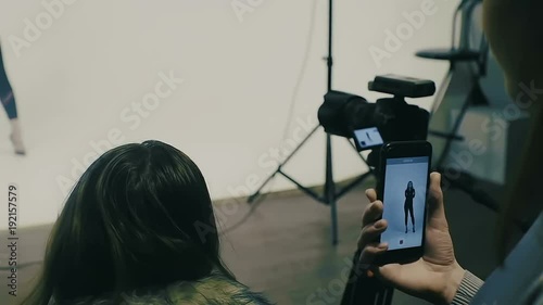 Young beautiful female model posing for a photo shoot in a studio, photographer is shooting with smartphone Slow Motion backstage