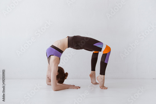 Beautiful young woman working out in white interior, doing yoga exercise, stretching, standing in Bridge Pose, Urdhva Dhanurasana (Upward Bow), Chakrasana (Wheel) Posture, full length. photo