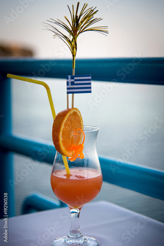 A glass with an orange and a cocktail and with a Greek flag stands on a table in a cafe in the evening.