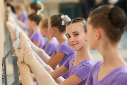 Beautiful smiling ballerina stretching leg. Young lovely ballerina training stretch of her leg and looking at camera close up. Kids ballet rehearsal.