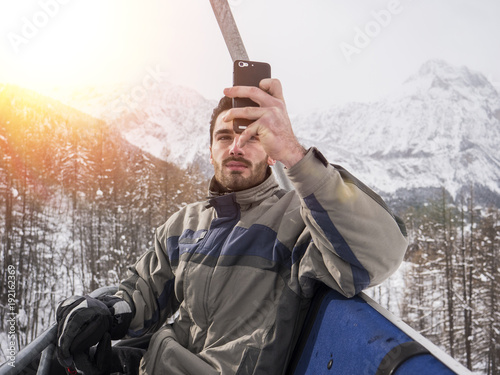 Handsome young man using smarpthone to take photographs of the landscape, while sitting on chairlift among snowy woods in the mountain