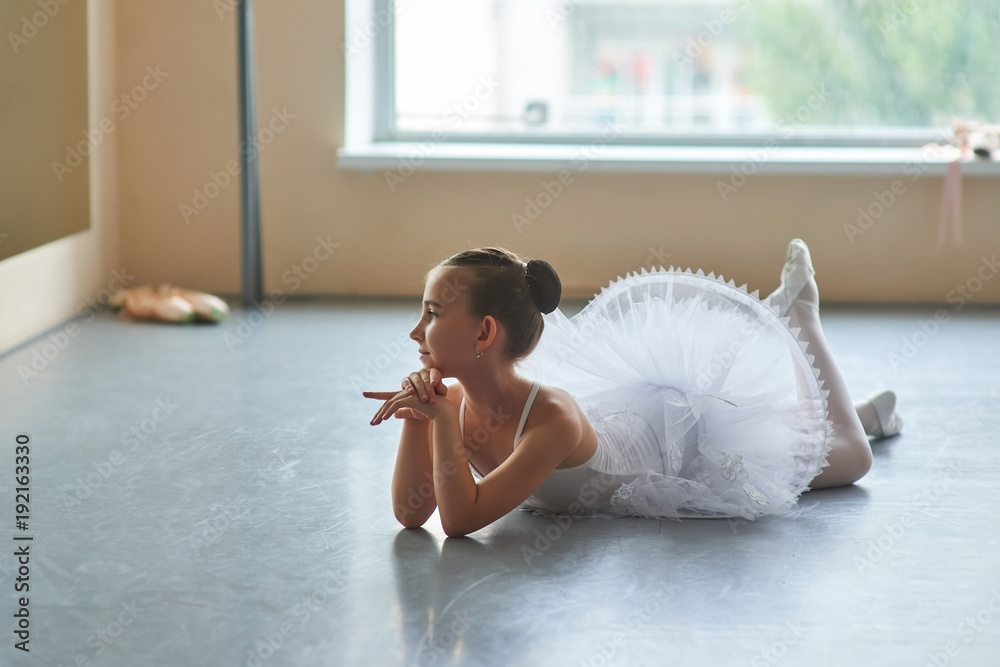 Obraz premium Adorable ballerina lying on the floor. Beautiful young ballet girl in white tutu posing on the floor in ballet class. Teenager ballerina on blurred background.