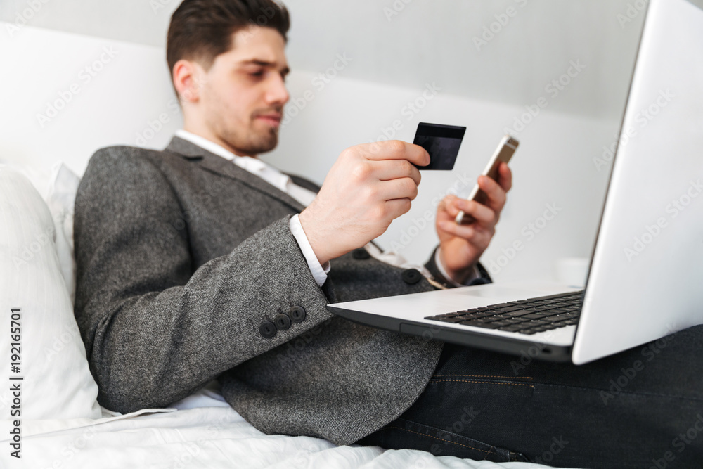 Photo of smart man in suit lying in bed, while paying online with credit card using laptop and cell phone Photo of smart man in suit lying in bed, while paying online with credit card using laptop and cell phone
