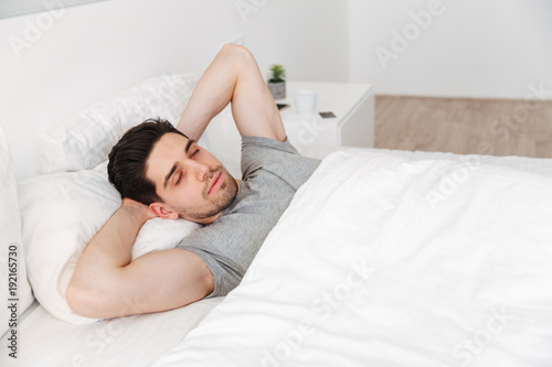 Portrait of sleepy unshaved man in casual t-shirt, having nap alone at home in bed with white clean linen putting hands under head