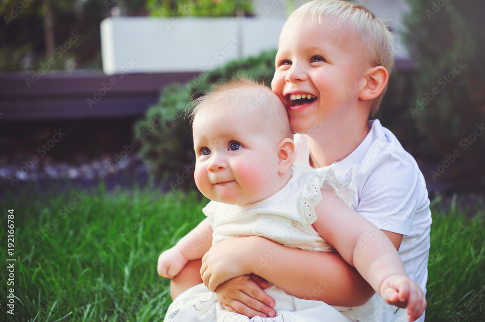 Fototapeta premium happy little brother playing hugs his sister baby sitting on grass in a green garden, concept of love and parenting