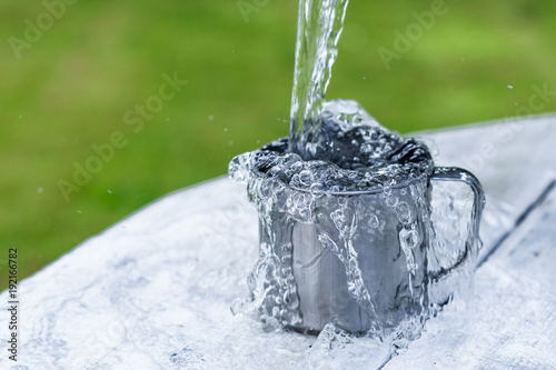 A steel mug with water and sprays on a white wooden table on nature background, life concept Water is pouring out of the bucket into the mug on a white wooden table on nature background