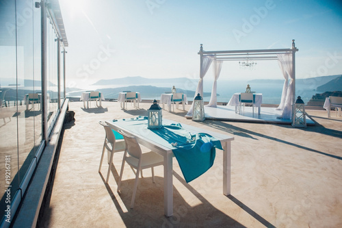 White arch for a wedding ceremony in the open air with lanterns and a platform with white furniture on the background of the sea, islands and blue sky on the island of Santorini