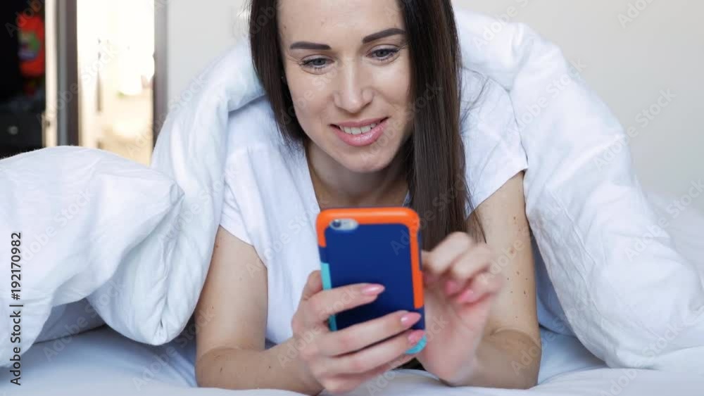 Smiling young female lying down the bed and using her phone Smiling young female lying down the bed and using her phone