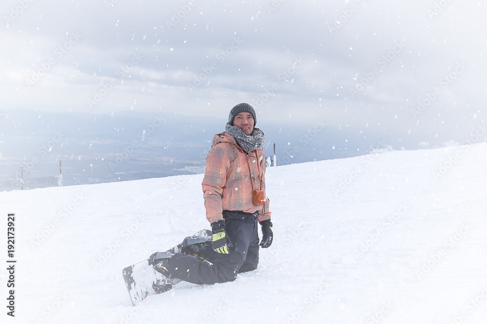 Portrait of snowboarder wearing sweater hat, orange & black stripe jacket, gloves and pants knees sitting  on top of mountain, looking at the camera on the background of hills. Snowflake, winter 