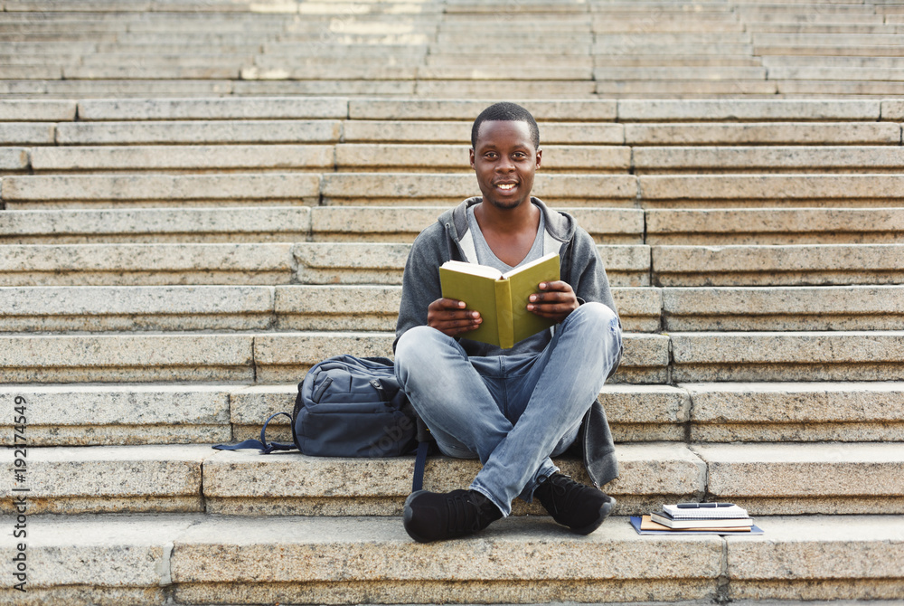 Smiling african-american student reading book on university stairs
