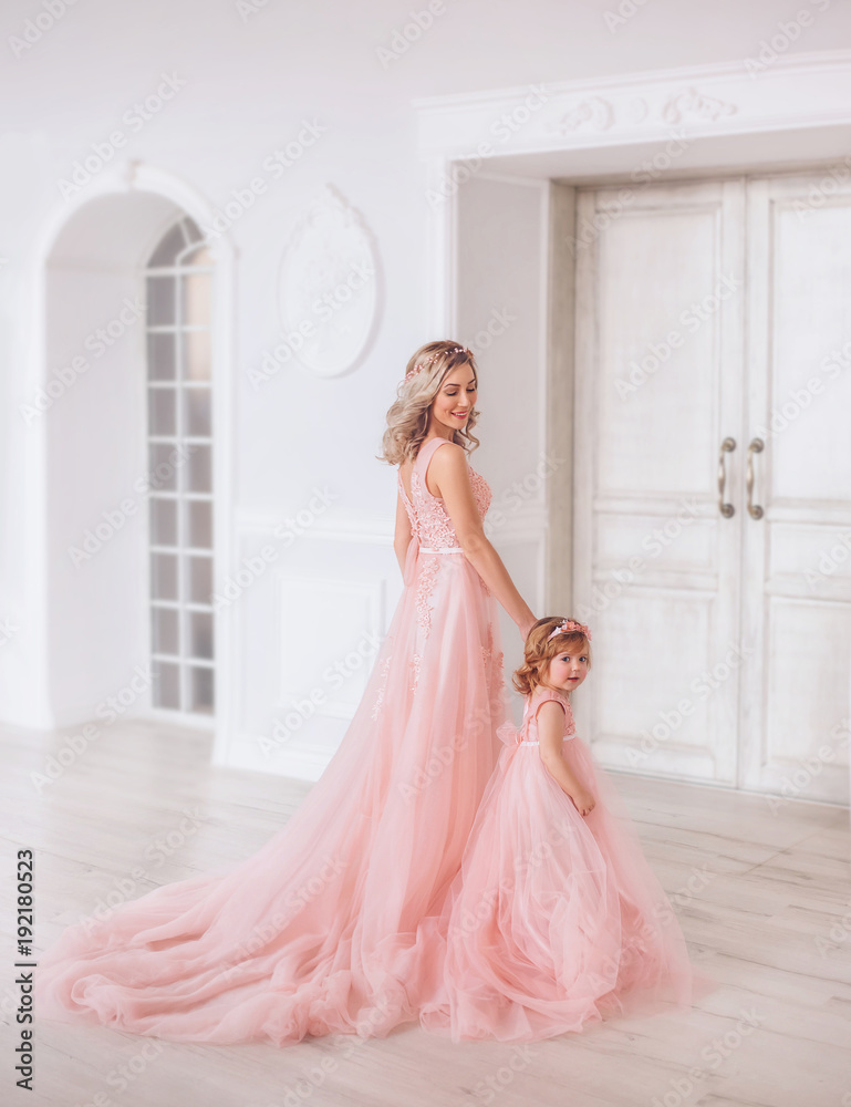 Mom and daughter in luxurious, pink dresses with a long train. Family clothes, identical dresses. The background is beautiful, expensive, white, classic interior. Artistic, family photography Mom and daughter in luxurious, pink dresses with a long train. Family clothes, identical dresses. The background is beautiful, expensive, white, classic interior. Artistic, family photography