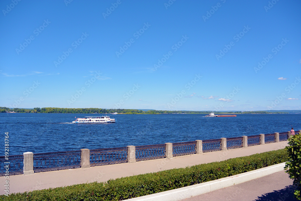 Obraz premium View on the Volga quay of the Samara city in sunny summer day. City embankment, barge and touristic motor ship boat on river, clear blue sky
