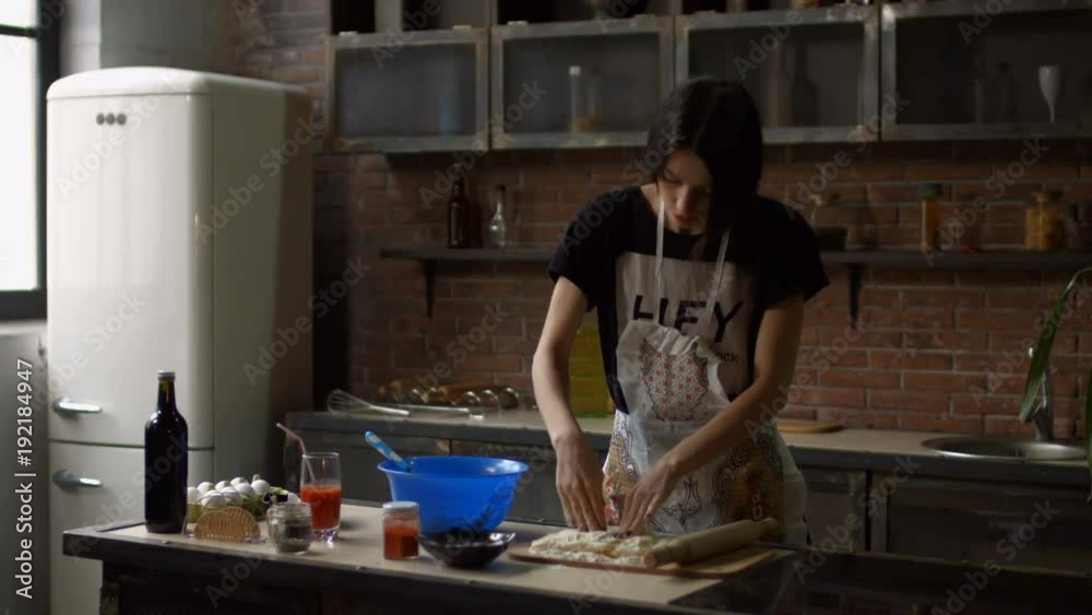 Attractive young brunette woman kneading dough in flour with hands while cooking in the kitchen. Cheerful housewife in apron rolling out dough with rolling pin in her home kitchen.
