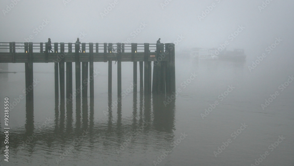 Silhouettes of people and bicycle on wooden pier on river Thames in London during dull gray foggy evening.