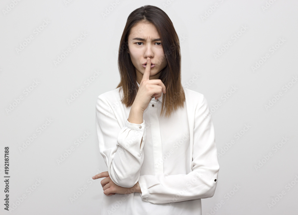 Portrait of a angry serious young woman with dark hair and white shirt, looking into the camera, showing silence sign, pressing her finger to her lips on the white background Portrait of a angry serious young woman with dark hair and white shirt, looking into the camera, showing silence sign, pressing her finger to her lips on the white background