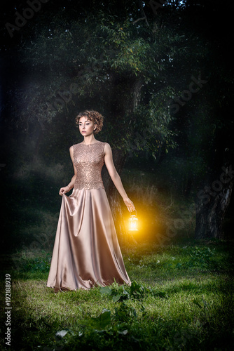 Art photo shoot. Beautiful sexy slim female model with short brown curly hair in wonderful gold long evening dress with lamps light, outdoor in fairytale dark green forest. Fantasy.