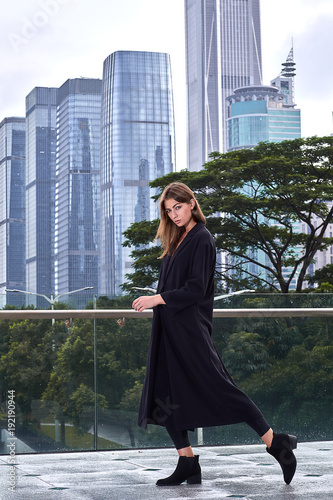 Young beautiful sexy woman with brunette hair in a long black coat, boots and casual style perfect posing near the skyscrapers in Hong Kong. Fashion street vogue model outdoor photo shooting