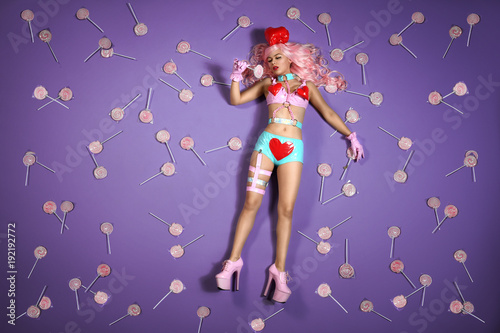 lebende Barbiepuppe in knappen blauen Lackhotpants und high heels liegt träumend inmitten von vielen Lollypops auf einem knallig Violetten Hintergrund.