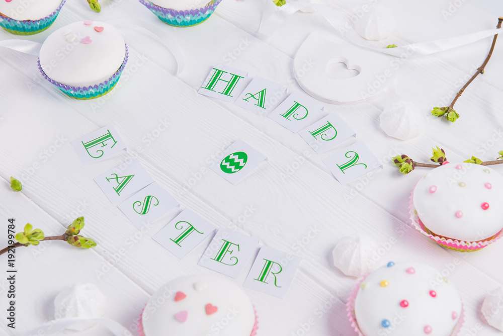 Holiday composition of Happy Easter lettering, branches with young shoots of greenery, decorated cupcakes, merengue sweets, bird figure on wooden background. Art concept. Selective focus.
