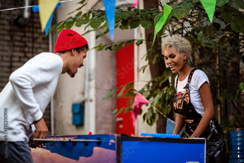 Two mixed race female and male friends play table football outdoor, entertain themselves. Positive stylish people play outdoor game together. People, recreation and entertainment concept. photo