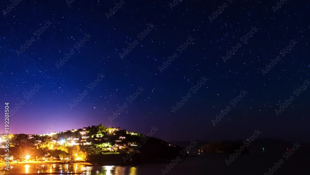Time lapse of a starry sky above a small city illuminated in front of the sea and a beach