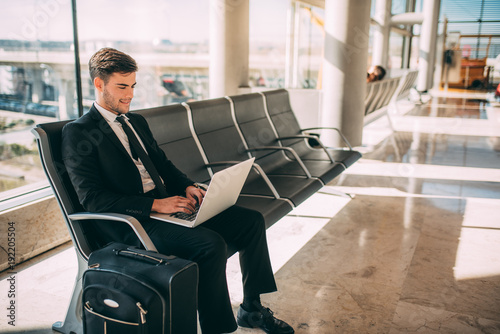 Young business man sitting on the computer with the suitcase at the airport waiting for the flight 